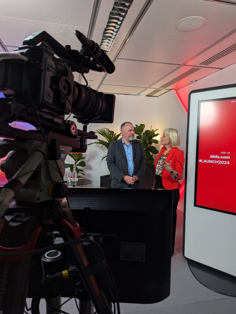 Kirsten wearing an orange jacket and a headset presenting next to a panel alongside a man, behind large broadcast cameras